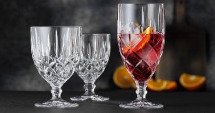 The NACHTMANN Noblesse iced beverage glass filled with a red cocktail drink, ice cubes and a slice of orange besides the empty tall and the empty small goblet. In the background are oranges.<br/>