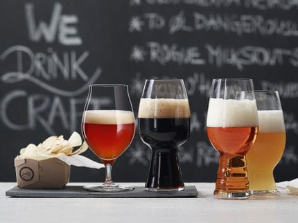 Les verres à bière artisanale SPIEGELAU remplis de bière sont posés sur une table devant un tableau noir avec un menu. Le verre à bière vieillie en fût et le verre à Stout sont posés sur une plaque d'ardoise, le verre à IPA et le verre à bière de blé américaine sont posés à côté sur la table.<br/>